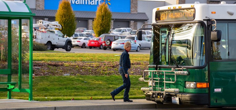 A passenger is approaching a green public transit bus at a bus stop, with the destination sign on the bus displaying '207 WALMART/YMCA'. The bus stop features a shelter and bench, and the background includes a Walmart store, indicating the stop is in a commercial area. The scene is set in daylight with clear skies and a parking lot full of cars behind the stop.